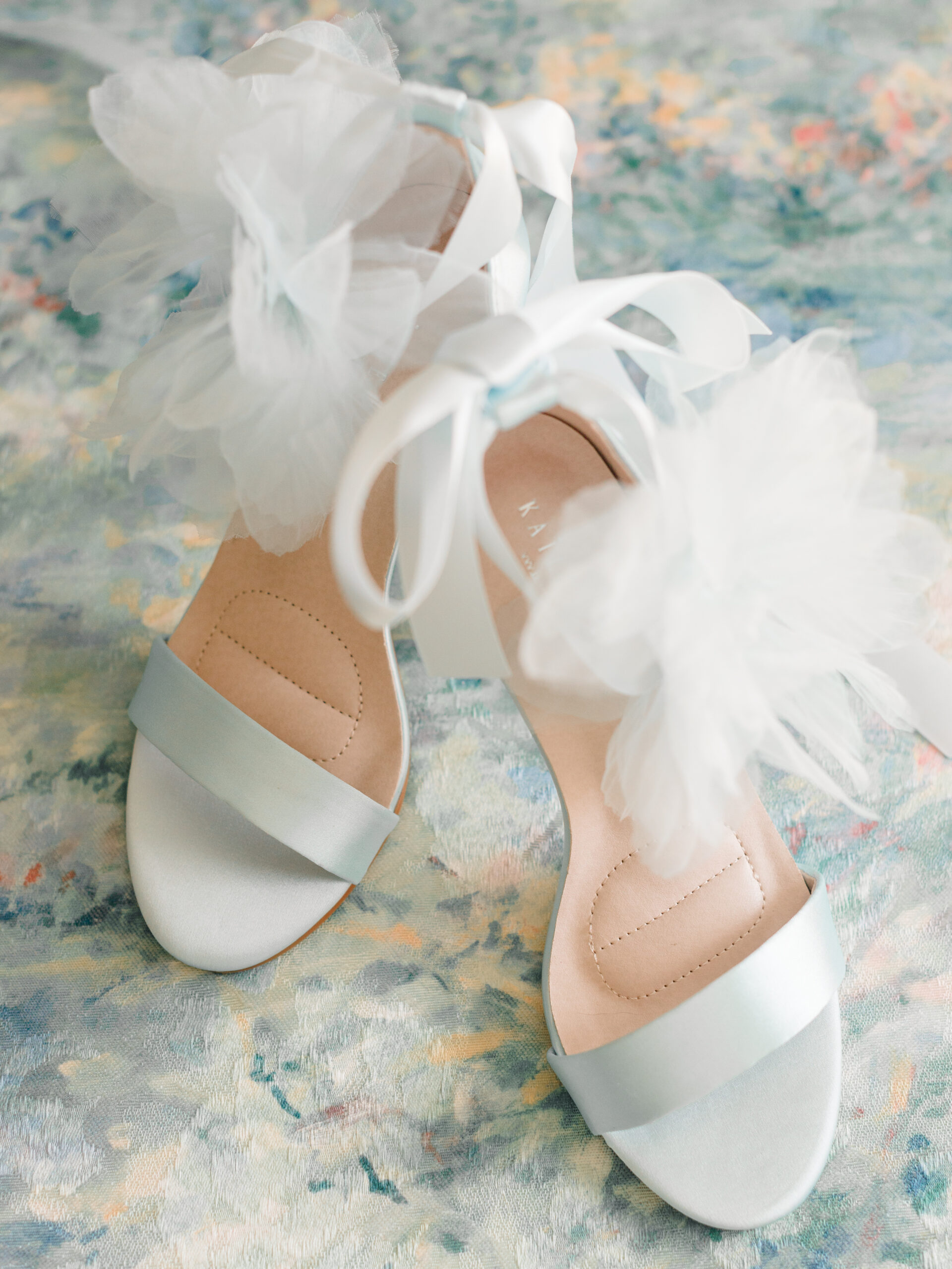 Kailee P wedding shoes in the Ophelia style, featuring satin straps and delicate tulle flower accents, styled on a painterly pastel backdrop for a romantic flatlay composition.