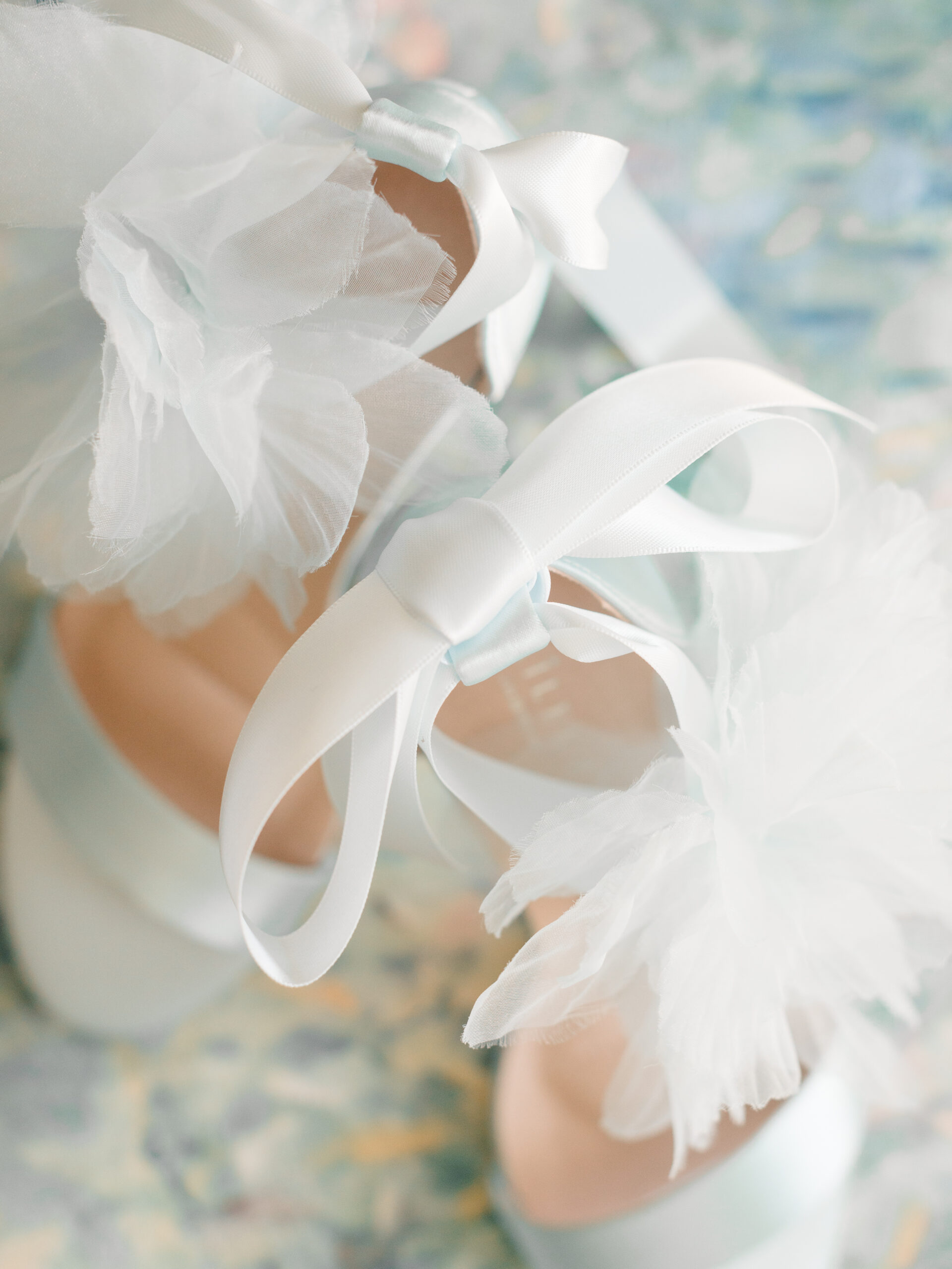 Close-up of Kailee P wedding shoes featuring the Ophelia design, highlighting the soft white ribbon ties and layered tulle floral accents on a painterly blue and green background.