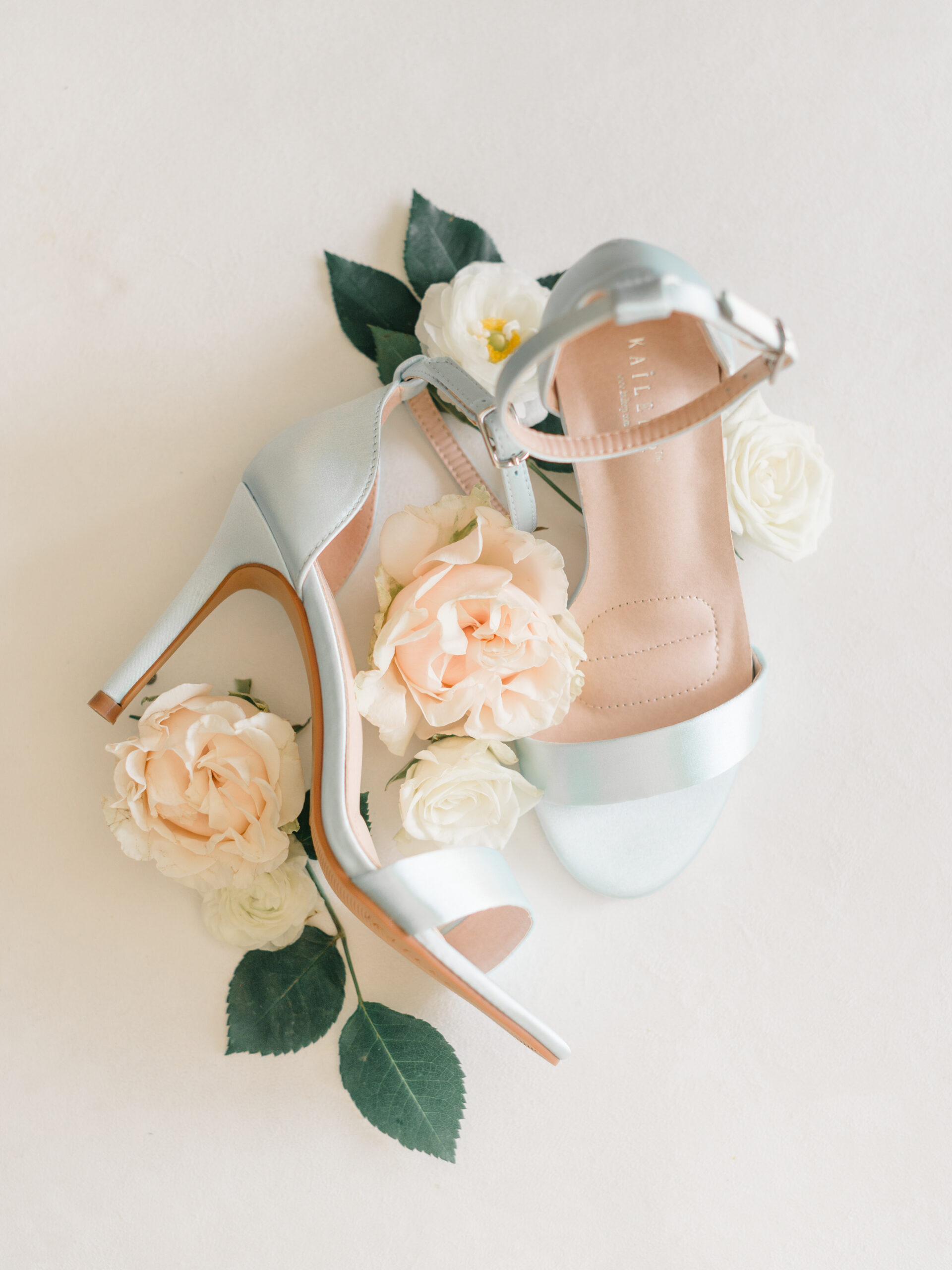 Flatlay of the Ophelia bridal heels from Kailee P wedding shoes, styled with ivory and blush garden roses on a neutral backdrop—showcasing the romantic satin finish and delicate ankle strap design.