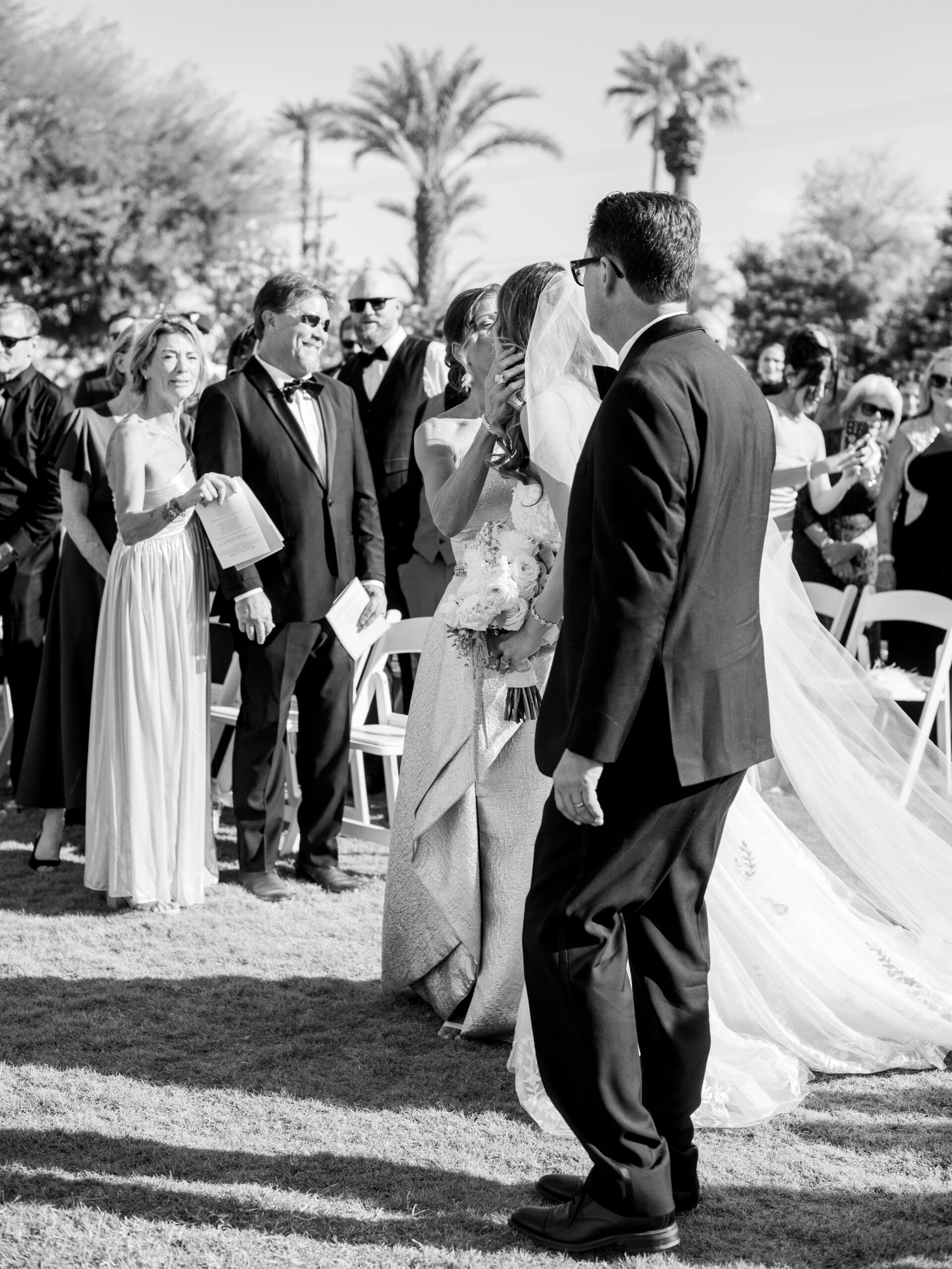 Emotional black-and-white photo of the bride embracing her parents during the wedding ceremony at Bougainvillea Estate in Indio, CA, surrounded by guests and palm trees.