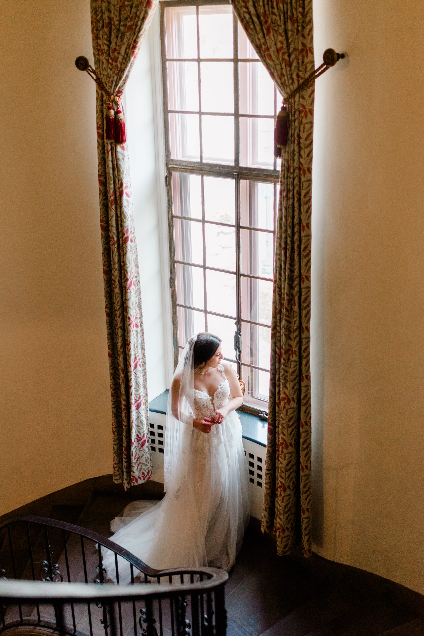 Bride standing by a tall arched window in an elegant historic staircase at Tyler Gardens, captured in soft natural light — a timeless moment from a bespoke wedding photography experience in Philadelphia by Elizabeth Kane Photography.