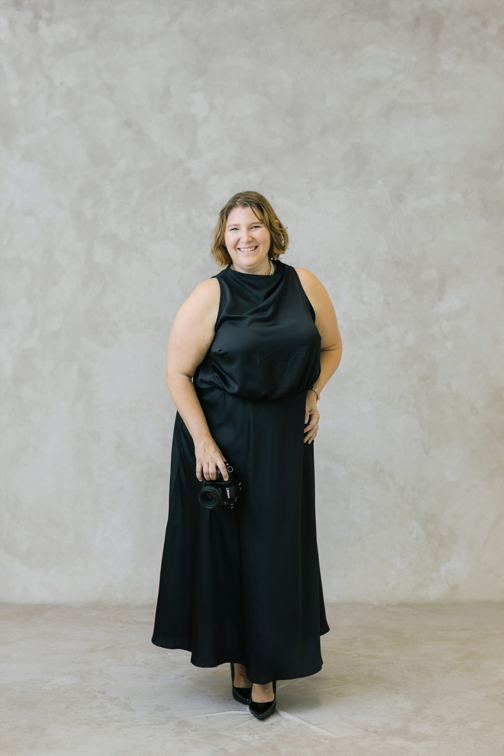 Portrait of Elizabeth Kane, Philadelphia fine art wedding photographer, wearing a black gown and holding a camera in a light-filled studio setting.