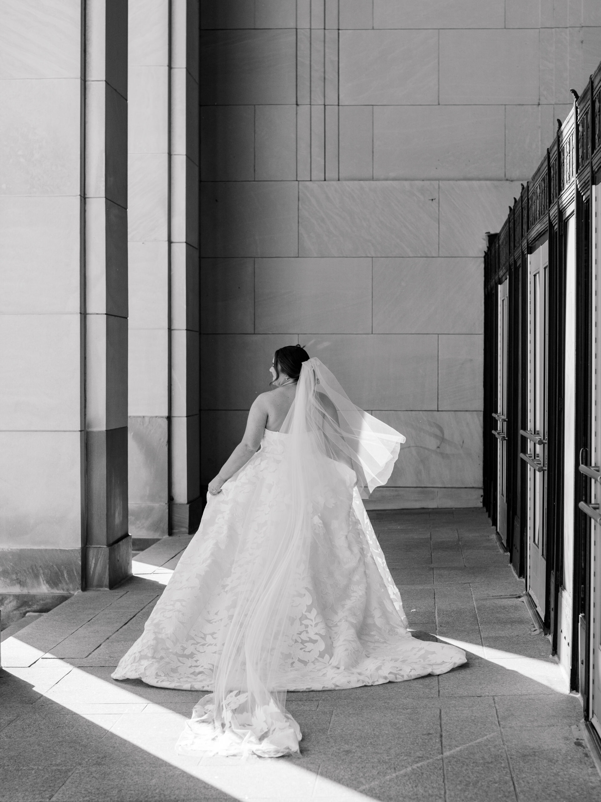 Bride in a strapless lace wedding gown walking through a sunlit stone corridor, captured in a timeless Philadelphia documentary wedding photography style.