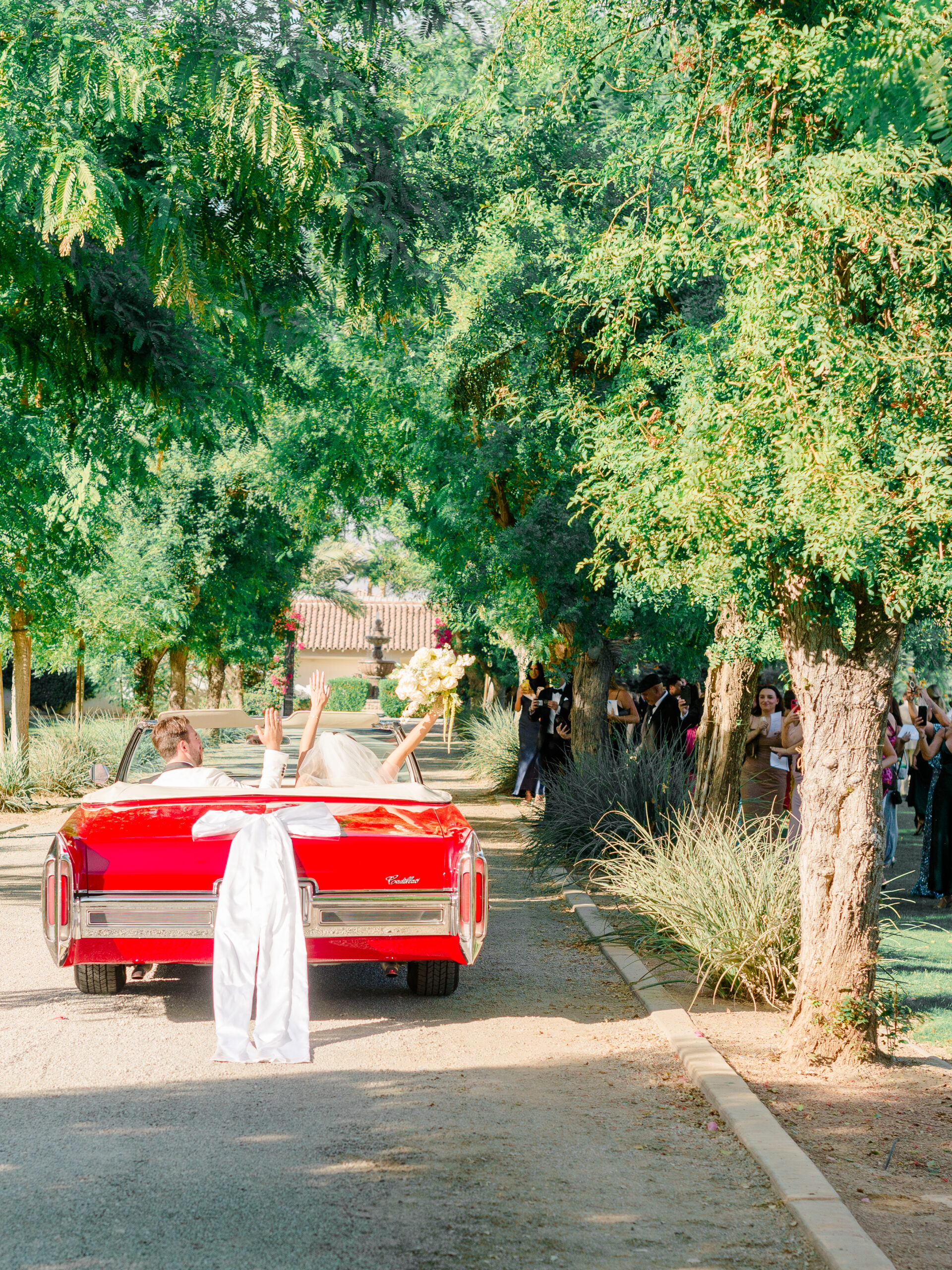 Bride and groom making a joyful exit in a vintage red Cadillac convertible, surrounded by cheering guests under a canopy of trees—captured by a documentary wedding photographer in Philadelphia.