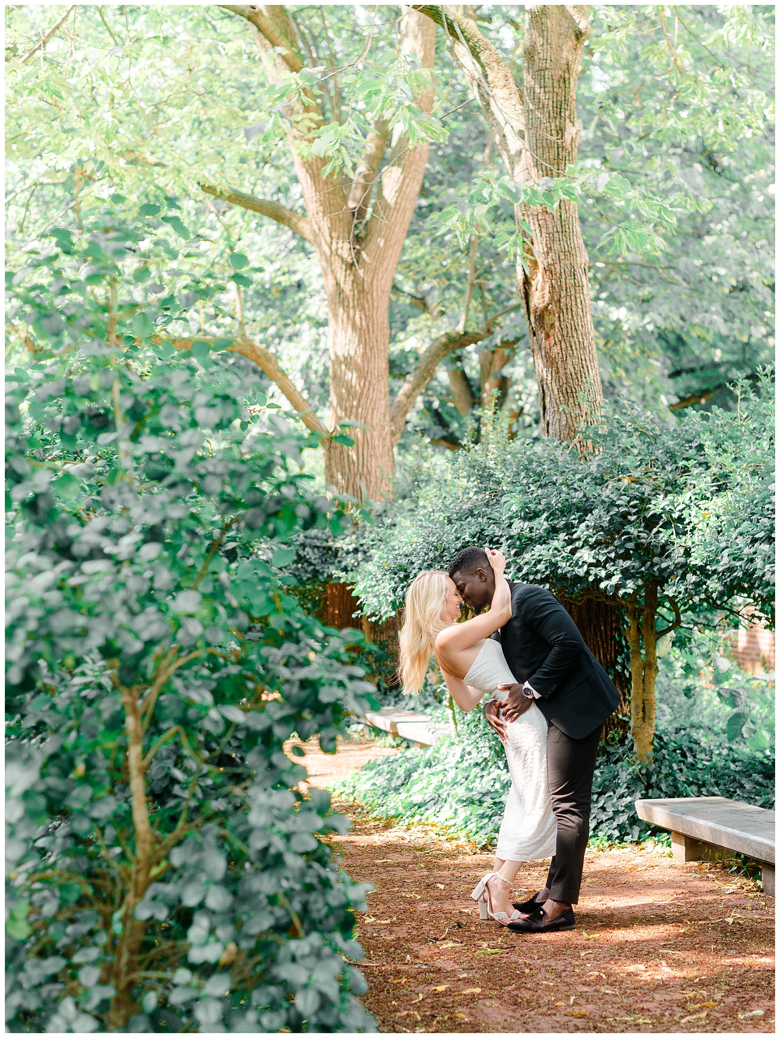 Romantic Old City Philadelphia engagement session with a couple embracing under lush greenery, the groom-to-be in a black suit dipping the bride-to-be in a strapless white dress for an elegant kiss.