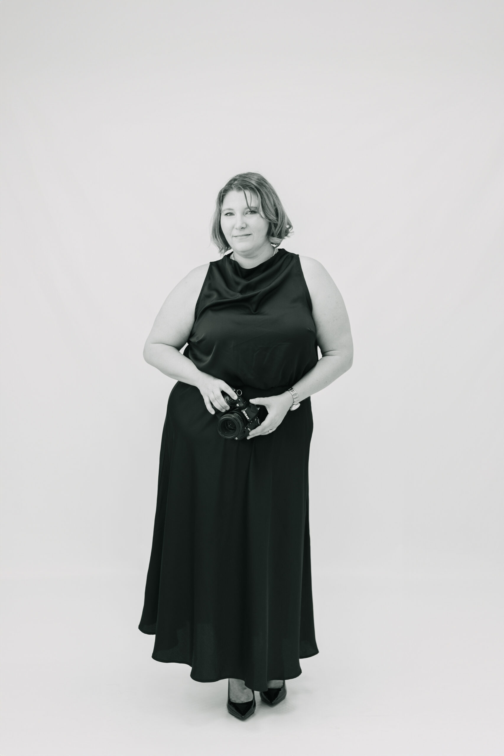Boutique full-service wedding photography represented by a refined black and white portrait of wedding photographer Elizabeth Kane holding her camera in a minimalist studio setting.