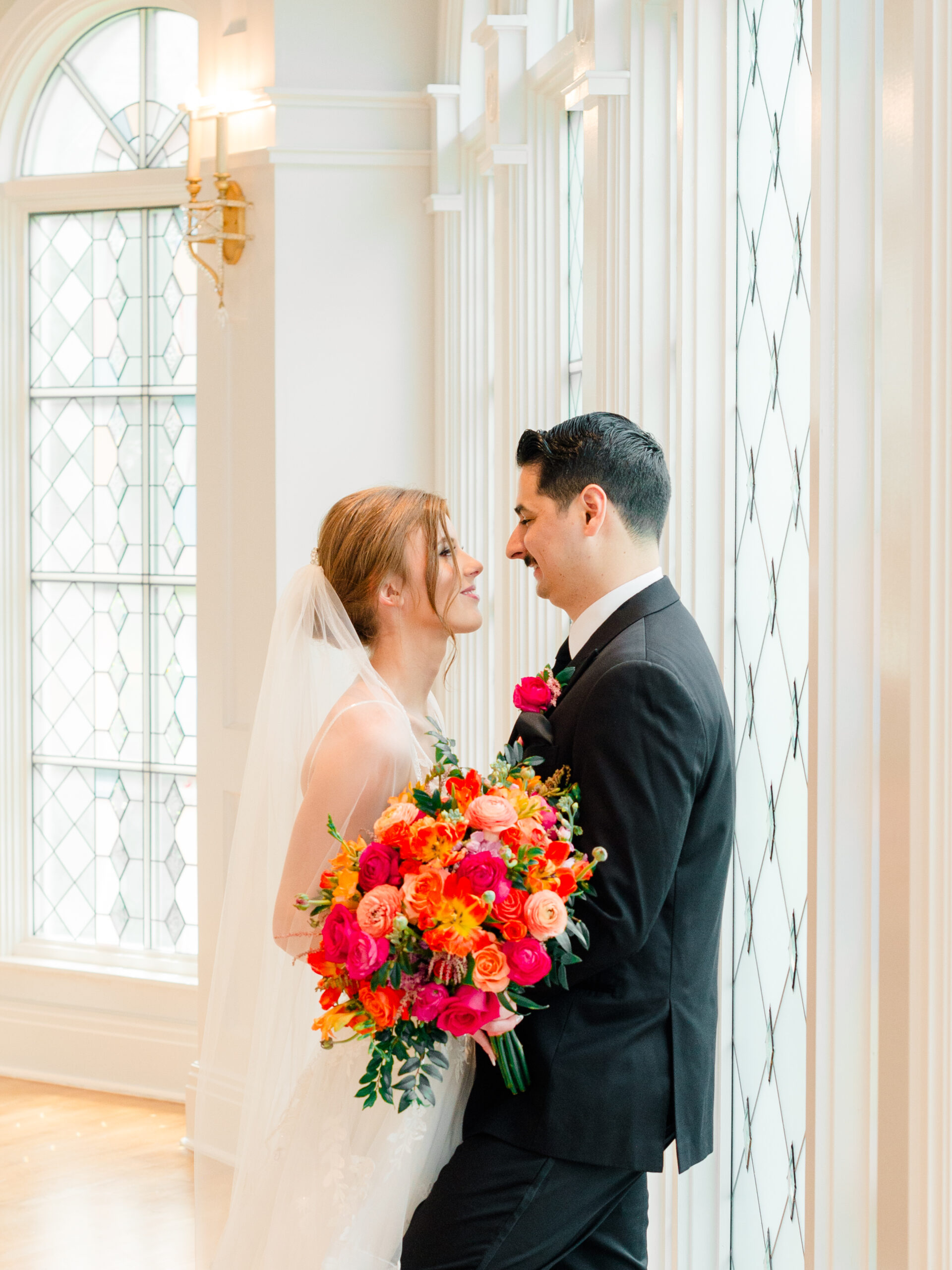 Boutique full-service wedding photography capturing an intimate indoor portrait of a bride and groom sharing a quiet moment beside tall windows with a vibrant floral bouquet.
